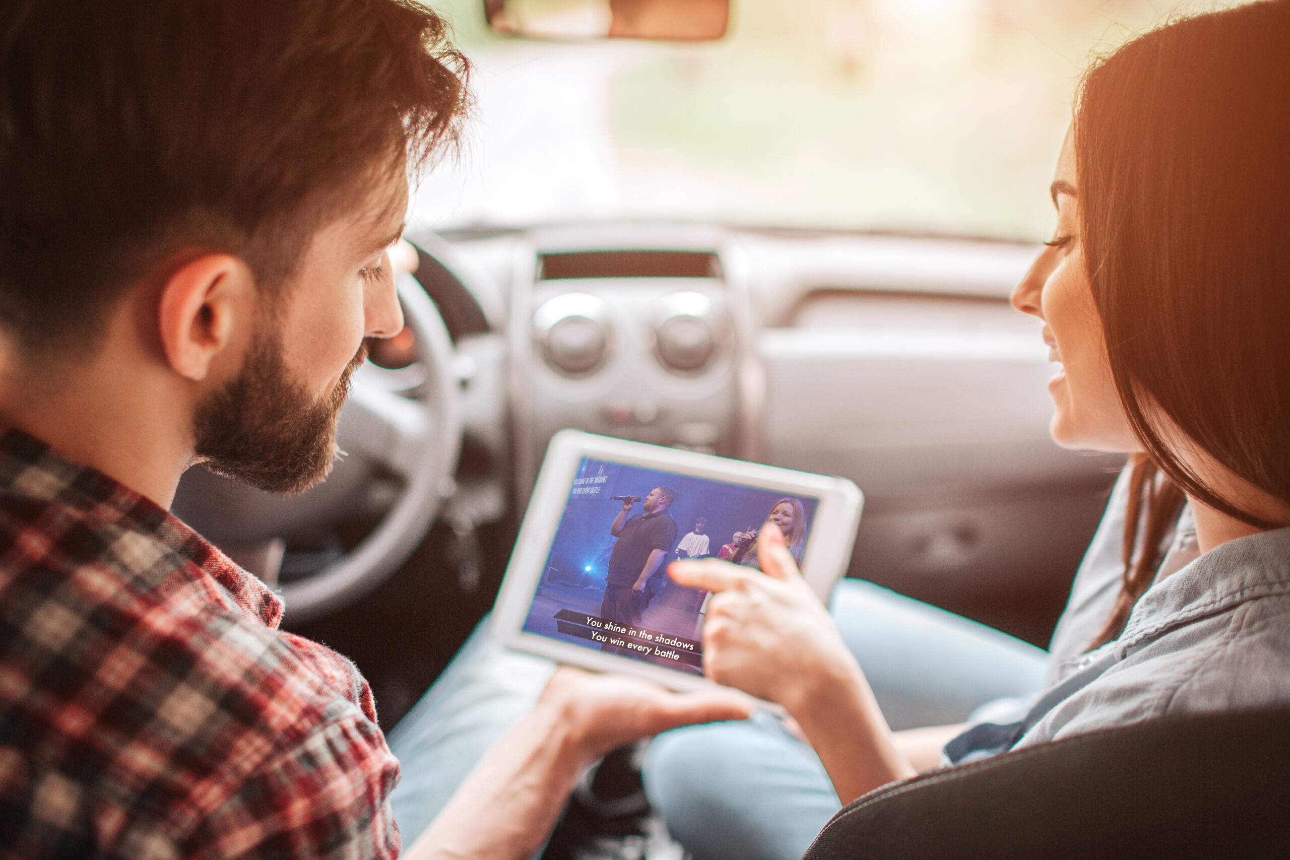 Guy is sitting in car with girl and looking at tablet with dark screen. He holds device in his hand. Girl is pointing on screen with her finger.